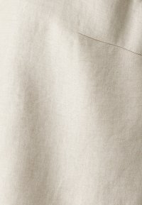 Beiger Baumwollstoff mit einem subtilen diagonalen Webmuster. Glatte Textur mit einer fast einheitlichen Oberfläche. Hat ein einzelnes gesticktes Detail.