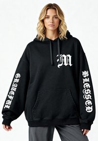Hoodie oversize nero, in cotone, con tasca frontale, cappuccio con cordino e testo bianco: "M" sul petto, "GRATEFUL" e "BLESSED" sulle maniche.