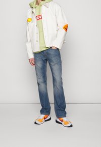 Veste blanche avec poches ornées d'appliqués colorés, sweat à capuche vert, jean bleu, et baskets multicolores avec des motifs audacieux.