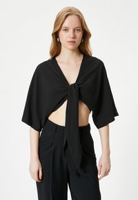 Sort crop top med en knudedetail foran, korte, vide ærmer og en dyb V-udskæring. Parret med højalvet sorte bukser.