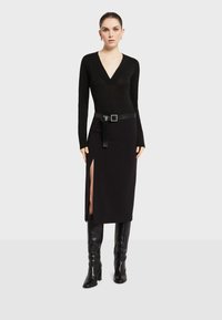 Crna priljubljena majica dugih rukava s V-izrezom, u kombinaciji s crnom midi suknjom koja ima bočni prorez i sjajnim remenom sa kvadratnom kopčom.