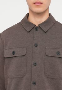 Άνδρας που φοράει καφέ πουκάμισο-σακάκι με δύο τσεπάκια στο στήθος με κουμπιά και κλασικό γιακά.