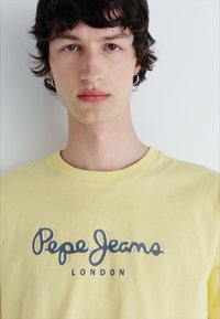 Geltona medvilninė marškinėliai su tamsiai mėlynu „Pepe Jeans London“ logotipu kreivais rašmenimis. Klasikinio apvalaus kaklo dizainas. Standartinio dydžio.
