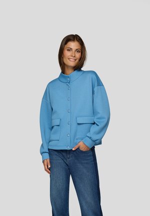 Blaues strukturiertes Jacket mit hohem Kragen, großen Fronttaschen und Knopfverschluss. Über einer Jeans getragen.