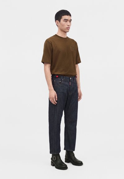 Homme debout portant un t-shirt marron à manches courtes, un jean en denim bleu foncé avec une ceinture noire, et des bottines noires à boucles.