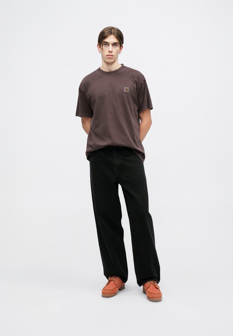 Braunes Baumwoll-T-Shirt mit einem kleinen Logo-Patch, kombiniert mit locker sitzenden schwarzen Hosen und orangenen Schuhen, steht vor einem einfarbigen Hintergrund.