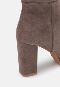 Botín de ante gris de tacón alto, con un tacón robusto, punta redondeada y textura suave, que muestra detalles de costura a lo largo de las costuras.