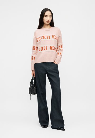 Γυναίκα με ροζ πουλόβερ με το κείμενο "Rock n Roll", σκούρο παντελόνι με φαρδές πιέτες, μαύρα μυτερά παπούτσια, κρατώντας μαύρη τσάντα στο αριστερό της χέρι.