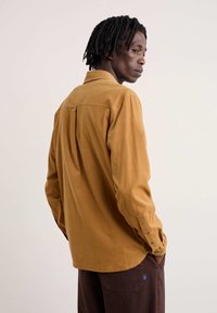 Een lichtgekleurde blouse met lange mouwen en knopen in mosterdkleur, gemaakt van textiel met structuur en een achterste plooi; gecombineerd met bruine broek met een logo.