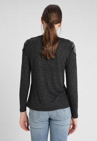 Chemise à manches longues gris foncé en tissu doux, avec des accents décoratifs sur les épaules et un ourlet courbé. Associée à un jean bleu clair.