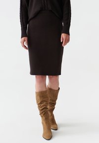 Jupe midi en tricot marron associée à des bottes montantes en suède beige. La tenue présente un tissu confortable et texturé avec une silhouette ajustée.