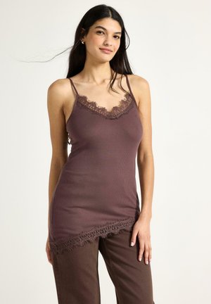 Donna che indossa un top aderente color malva con rifiniture in pizzo e pantaloni marroni, in piedi davanti a uno sfondo chiaro uniforme.