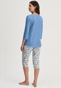 Lichtblauw, langemouwen top gecombineerd met witte, met bloemenpatroon bedekte capri-broek. Het ontwerp bevat kleurrijke bloemen in tinten blauw, groen en paars.