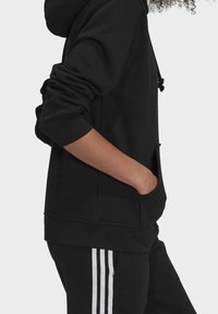 Person klädd i svart hoodie och svarta byxor med vita ränder, med handen instoppad i huvan, sedd från sidan.