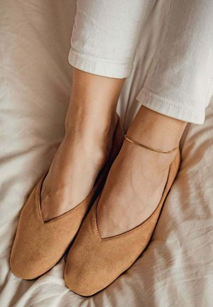 Ballerines en suède beige avec une ouverture en forme de V, texture douce et semelle flexible. Un bracelet fin en or à la cheville ajoute une touche subtile.