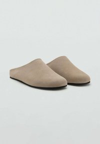 Pantofole beige senza lacci in morbido suede, con un design minimalista caratterizzato da una texture liscia e una suola nera flessibile.