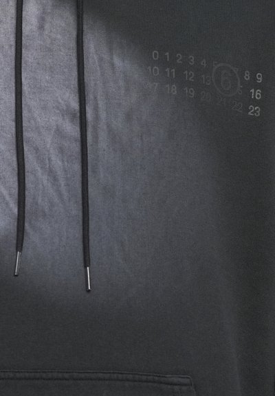 Črna kapuca z gladko teksturo, opremljena z dvema vrvicama z kovinskimi konicami in subtilnim tiskom številk na strani.