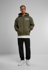 Felpa verde con zip, tasche frontali e dettaglio grafico blu, abbinata a pantaloni neri e sneakers bianche. Materiale in cotone liscio.