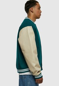 Jachetă varsity teal cu mâneci din piele bej. Prezintă un guler și manșete ribbate cu dungi, buzunare laterale și un design lejer.