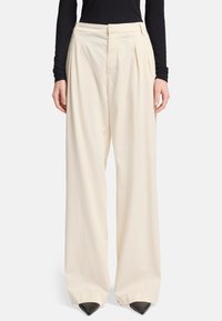 Crème-kleurige wijde broek met voorplooien, gemaakt van lichtgewicht stof, met een hoge taille en een ingestopte tailleband. Zwarte puntige schoenen.