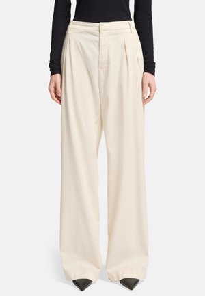 Crème-kleurige wijde broek met voorplooien, gemaakt van lichtgewicht stof, met een hoge taille en een ingestopte tailleband. Zwarte puntige schoenen.