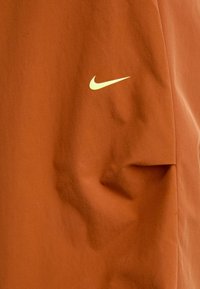 Nike športna oblačila v toplem oranžnem odtenku z gladko teksturo. Na sprednji strani se nahaja majhna rumena swoosh logo.