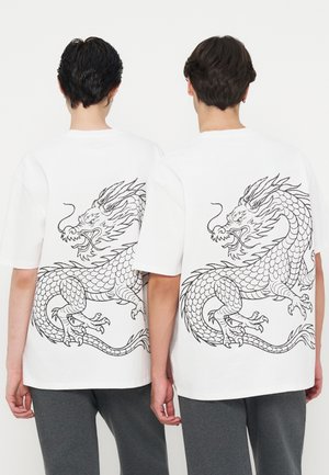 Deux personnes portant des t-shirts blancs avec de grandes illustrations noires de dragons dans le dos, debout côte à côte, dos à nous.