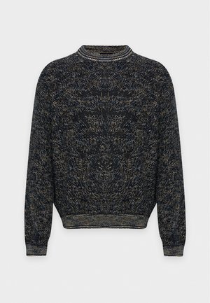 Un homme pose avec un pull foncé à la texture marquée, un pantalon cargo gris anthracite et des chaussures noires sur un fond clair et uni.