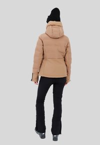 Chaqueta de plumas color marrón claro con capucha, con un diseño acolchado y detalles en negro, combinada con pantalones negros ajustados y botas de nieve con Cremalleras.