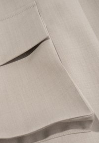 Beige stof met een gladde, gestructureerde oppervlakte, met een gevouwen pocketdetail. Het materiaal lijkt zacht, met subtiele stiknaden.