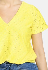 Dzeltena īsām piedurknēm blūze ar V veida izgriezumu, ar acs rakstu un ģeometriskām formām, apvienota ar džinsu apakšdaļām.