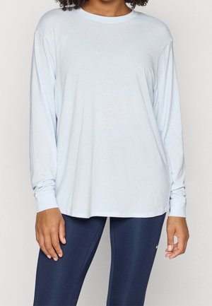 Osoba ubrana w luźną, jasnoniebieską koszulę z długim rękawem i ciemnoniebieskie legginsy sportowe stojąca na jednolitym tle.