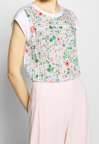 Top blanco sin mangas con un colorido estampado de flores y hojas, metido dentro de un pantalón de talle alto color rosa pastel, usado por una persona de pie.