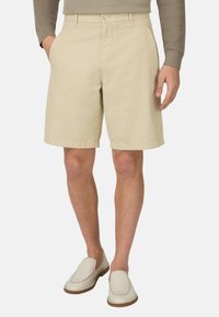 Homme portant un short beige jusqu'aux genoux, un haut à manches longues marron clair et des chaussures blanches sans lacets, debout les mains le long du corps.