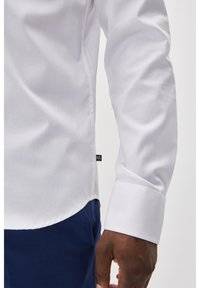 Witte langemouwen shirt met een gladde textuur, met een detail van de mouwopening en een klein zwart logo-label, gecombineerd met marineblauwe broeken.