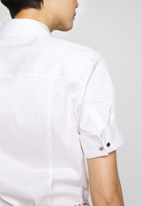 Wit overhemd met korte mouwen en een opstaande kraag, voorzien van opgerolde mouwen die zijn vastgezet met zilveren knopen en een glad, gladde stof zonder structuur.