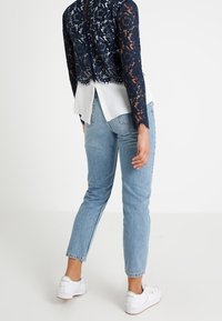 Person klädd i en marinblå spets långärmad topp, vit skjorta under, ljusblå jeans och vita sneakers som står på ett vitt golv.