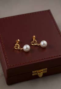 Guldörhängen i hjärtformat med en pärlstav, placerade på en mörkröd läderlåda med guldfärgad hårdvara och syddetaljer.