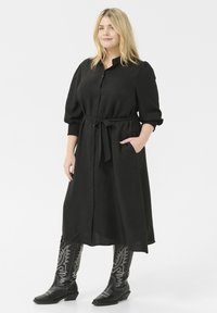 Black button-up dress with a tie belt and pockets, featuring three-quarter sleeves and textured fabric. Black embellished cowboy boots.