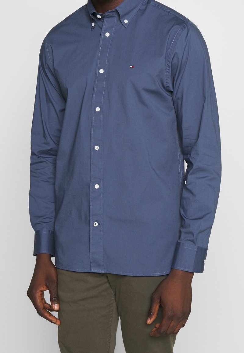 Camicia button-up in tessuto blu navy, maniche lunghe, colletto a punta, bottoni bianchi e un piccolo logo sul petto.