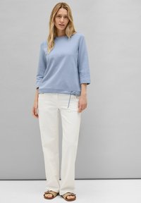 Un pull bleu clair texturé avec des manches trois-quarts et un lien ajustable à l'ourlet, associé à un pantalon blanc à jambes droites et des sandales marron.