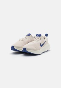 Chaussures de sport beige clair avec une tige en maille texturée, des accents bleus et une semelle coussinée avec une semelle extérieure en caoutchouc à motif.