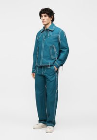 Άνδρας που φοράει τζελ μπουφάν και παντελόνι με λευκές ξεφτισμένες ραφές, στέκεται με τα χέρια στις τσέπες μπροστά σε απλό φόντο.