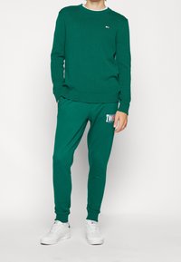 Zielony sweter z dzianiny z prążkowanym dekoltem i mankietami, w połączeniu z dopasowanymi dresami z logotypem. Buty są białe, co dodaje kontrastu.
