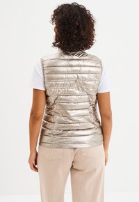 Metaliczna beżowa puchowa kamizelka z poziomym przeszyciem, bez rękawów i błyszczącą teksturą, noszona na białej koszuli i jasnych spodniach.