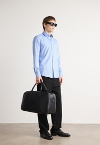 Bolsa de viaje de cuero negro con asas dobles, textura suave y herrajes plateados. Camisa de botones azul claro, pantalones negros y zapatos.