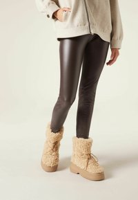 Tan fuzzy enkelboots met een gestructureerde buitenkant, ronde neus en platte rubberen zool; gecombineerd met donkerbruine nepleren leggings en een lichtgrijze zip-up jas.