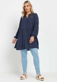 Navy button-up tunic with lace detailing, long sleeves, and gathered waist over light blue fitted jeans; paired with brown wedge sandals.