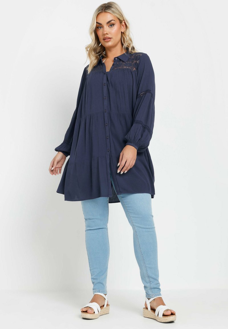 Navy button-up tunic with lace detailing, long sleeves, and gathered waist over light blue fitted jeans; paired with brown wedge sandals.