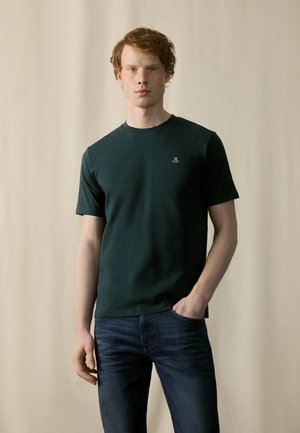 Dark green short-sleeve t-shirt with round neckline, featuring a small white logo on the left chest. Paired with blue jeans.